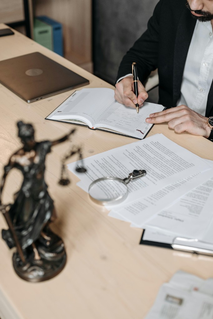 Professional in a blazer writing in a notebook at a desk with a Lady Justice statue, legal papers, a magnifying glass, and a laptop.