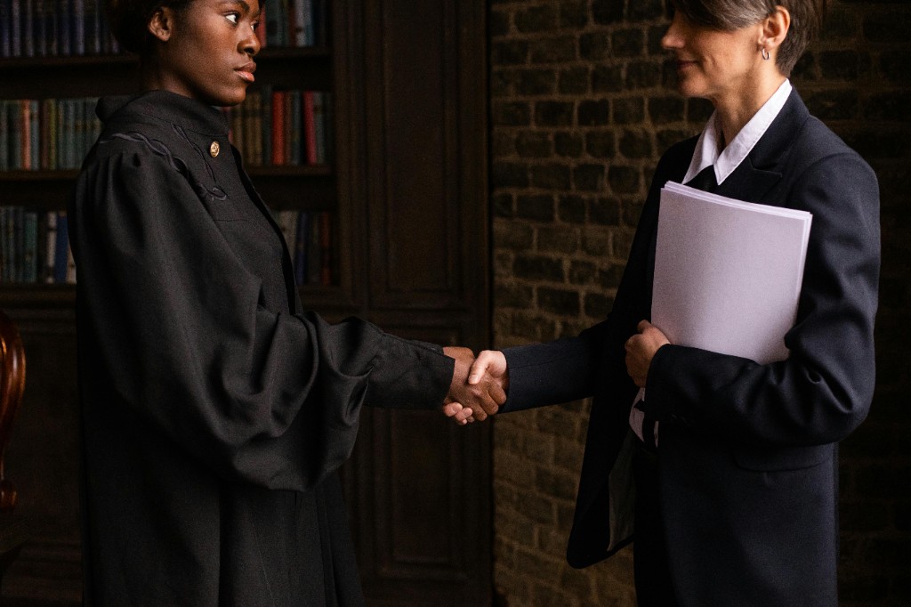 Two professionals shaking hands in a formal setting: one in an academic robe, one in a business suit holding papers, with law-library shelving in the background.