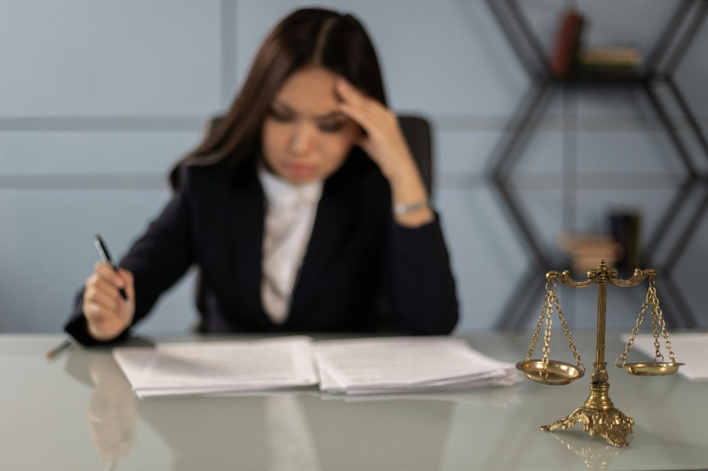Gold scales of justice in sharp focus on a desk; a professional in business attire works with papers in the softly blurred background.