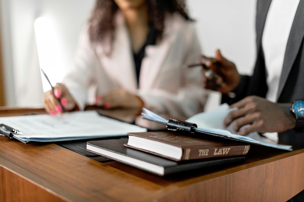 Two professionals collaborating over documents at a wooden desk with law books, including a volume titled “THE LAW” on the spine.