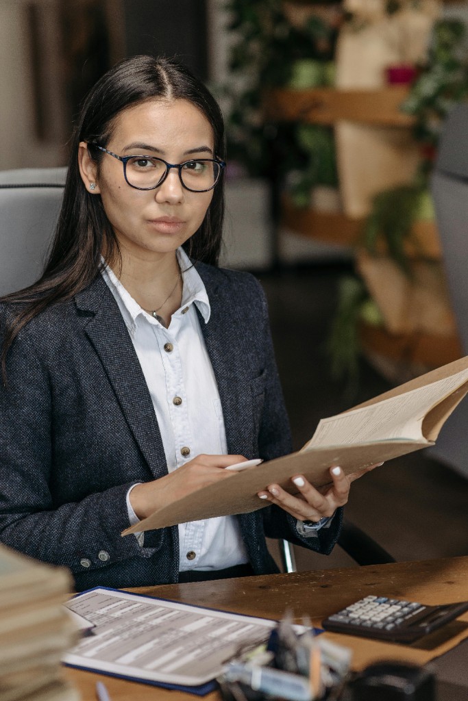 Professional woman in glasses and blazer reviewing documents at a desk in a bright office with plants and shelving.
