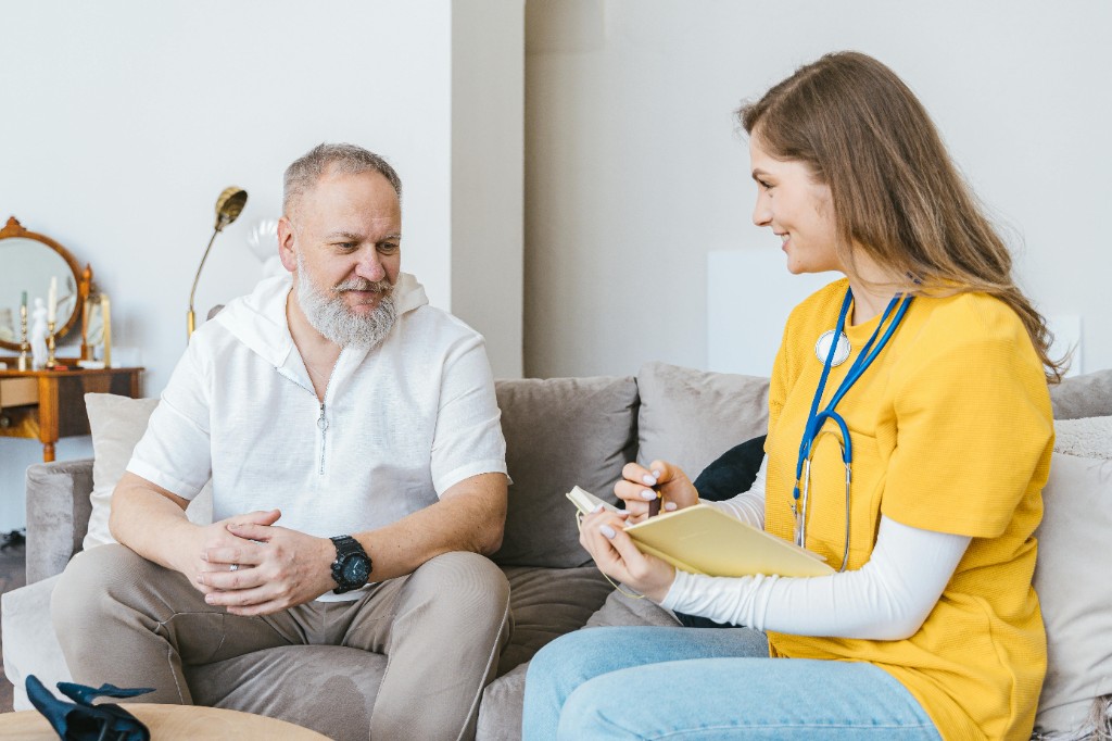 Healthcare worker wearing scrubs and a stethoscope speaks with an older adult seated on a couch, taking notes during a supportive conversation.