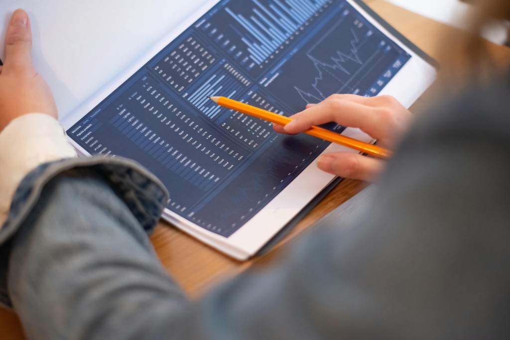 Hands using a pencil to point at data and charts displayed on a digital tablet screen, suggesting data analysis and reporting work.